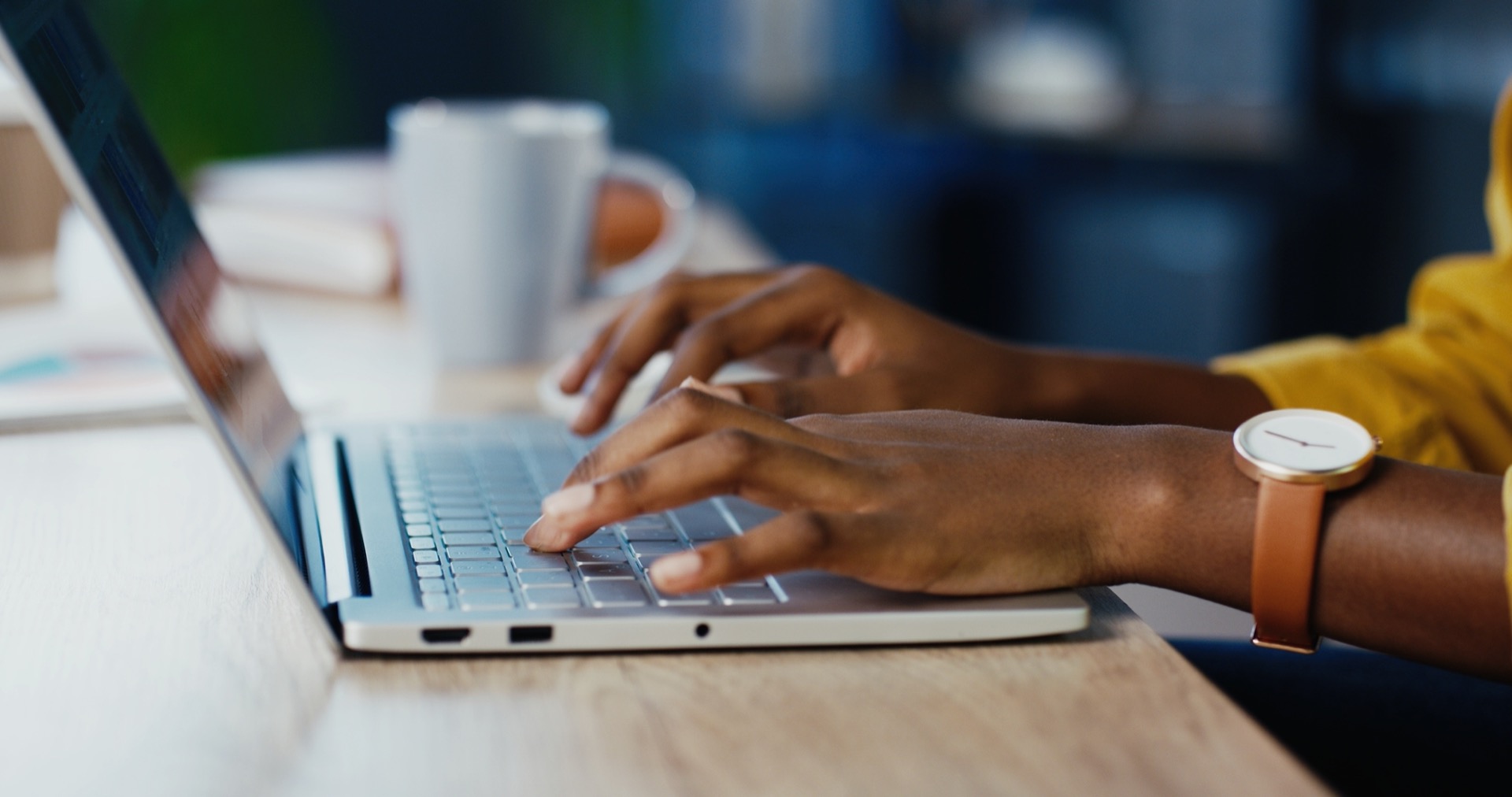 A woman with brown skin in a yellow top typing on a laptop at a wood desk &mdash; a small business owner taking the first steps to build her email list